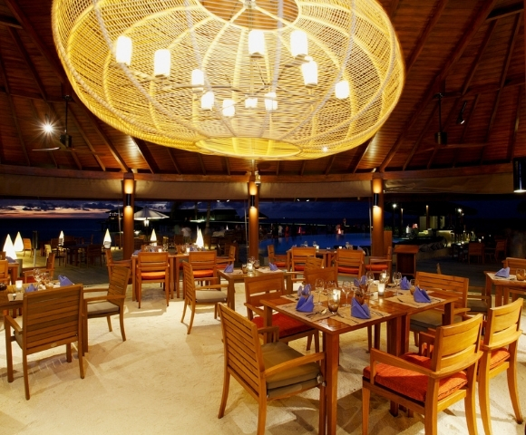 Elegant outdoor restaurant with wooden tables, vibrant blue napkins, and a large woven chandelier, illuminated against a night sky.