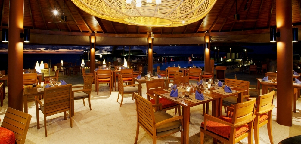 Elegant outdoor restaurant with wooden tables, vibrant blue napkins, and a large woven chandelier, illuminated against a night sky.