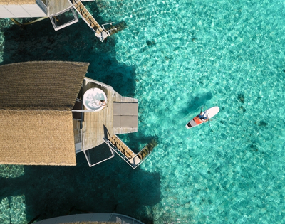 Aerial view of a thatched-roof overwater bungalow with a paddleboarder on vibrant turquoise waters.