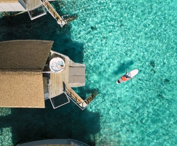 Aerial view of a thatched-roof overwater bungalow with a paddleboarder on vibrant turquoise waters.