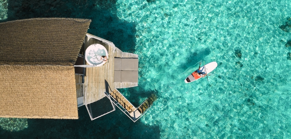 Aerial view of a thatched-roof overwater bungalow with a paddleboarder on vibrant turquoise waters.