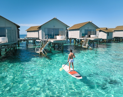 A person paddleboarding in clear turquoise water, surrounded by elegant overwater bungalows under a bright blue sky.