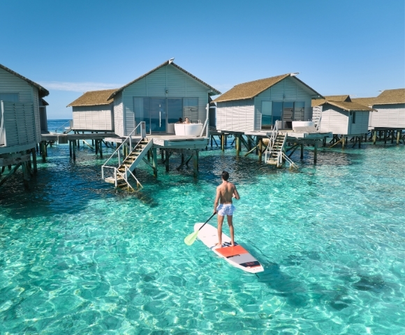 A person paddleboarding in clear turquoise water, surrounded by elegant overwater bungalows under a bright blue sky.