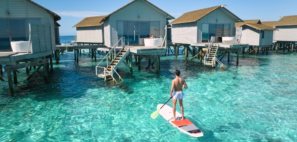 A person paddleboarding in clear turquoise water, surrounded by elegant overwater bungalows under a bright blue sky.
