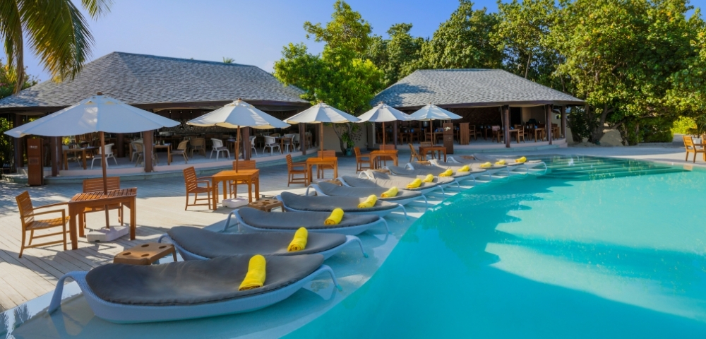 Serene poolside scene with loungers and umbrellas, surrounded by tropical foliage and a modern restaurant in the background.