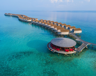 Aerial view of a luxurious overwater resort with thatched-roof bungalows and a circular lounge surrounded by crystal-clear turquoise waters.