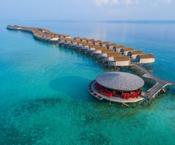 Aerial view of a luxurious overwater resort with thatched-roof bungalows and a circular lounge surrounded by crystal-clear turquoise waters.
