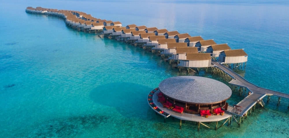 Aerial view of a luxurious overwater resort with thatched-roof bungalows and a circular lounge surrounded by crystal-clear turquoise waters.