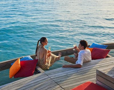 A couple enjoys drinks on a wooden deck by the water, surrounded by colorful cushions, with a calm ocean in the background.