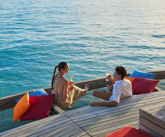 A couple enjoys drinks on a wooden deck by the water, surrounded by colorful cushions, with a calm ocean in the background.