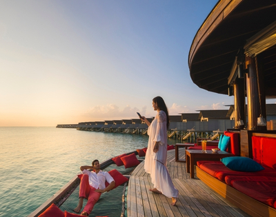 A couple enjoys a sunset on a wooden deck by the water, with one sitting relaxed while the other stands, capturing the moment on a phone.