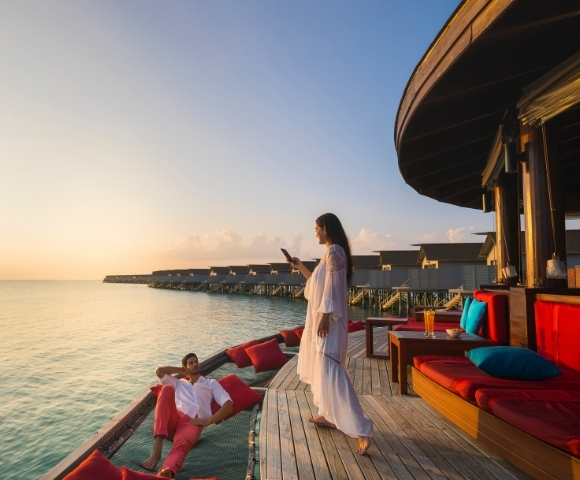 A couple enjoys a sunset on a wooden deck by the water, with one sitting relaxed while the other stands, capturing the moment on a phone.