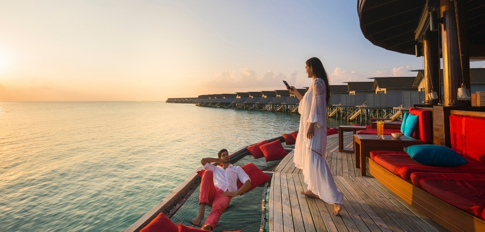 A couple enjoys a sunset on a wooden deck by the water, with one sitting relaxed while the other stands, capturing the moment on a phone.