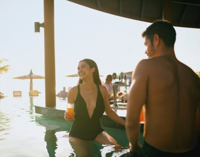 A woman in a black swimsuit sits in a pool at sunset, enjoying a drink, surrounded by relaxing vacationers and beautiful scenery.