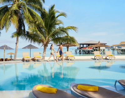 A couple strolls hand-in-hand by a tranquil pool with palm trees, tropical lounge chairs, and a seaside deck in the background.