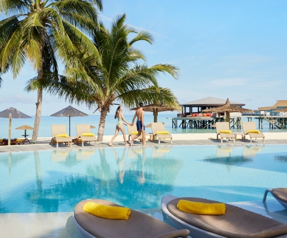 A couple strolls hand-in-hand by a tranquil pool with palm trees, tropical lounge chairs, and a seaside deck in the background.