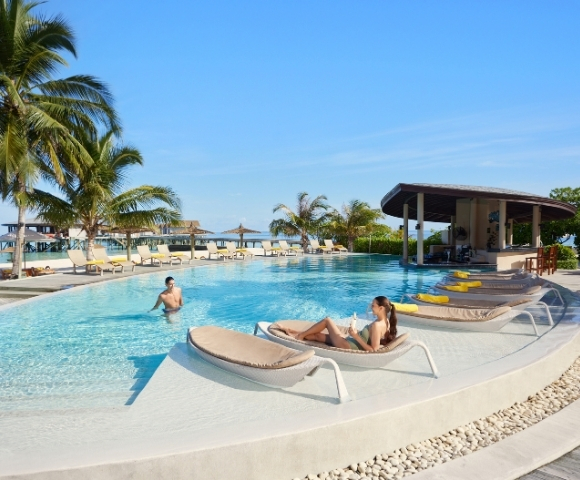 A serene poolside scene featuring a woman relaxing on a lounge chair and a man swimming, surrounded by palm trees and sunny skies.