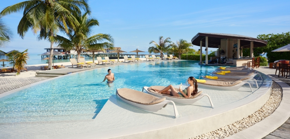 A serene poolside scene featuring a woman relaxing on a lounge chair and a man swimming, surrounded by palm trees and sunny skies.