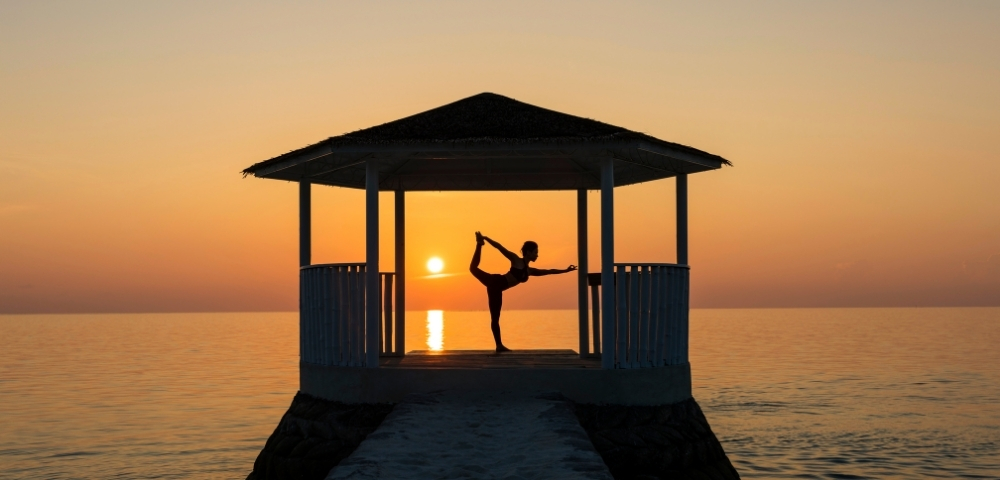 Silhouette of a person practicing yoga in a pavilion by the sea at sunset, creating a serene and tranquil atmosphere.