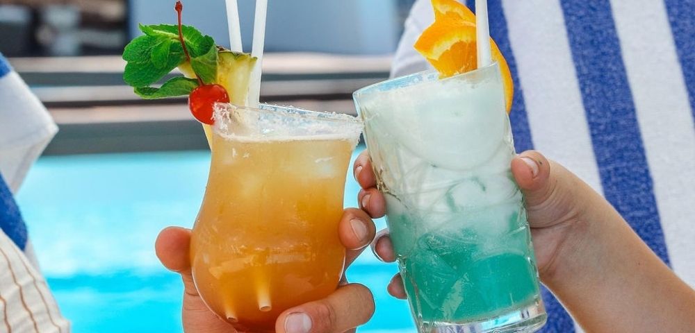 Two colorful cocktails are clinked together by hands, with a blue pool in the background, evoking a fun summer vibe.
