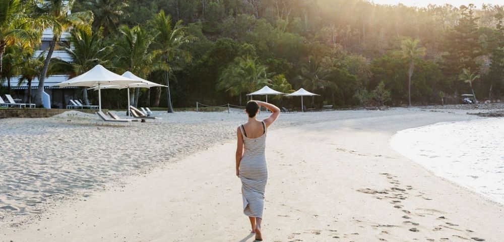 A woman in a flowing gray dress walks along a serene beach at sunset, palm trees and cabanas in the background.