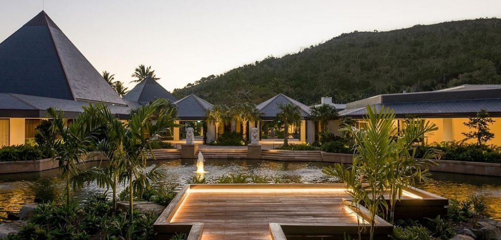 A serene resort at dusk, featuring a wooden walkway, lush greenery, and modern architecture set against a mountainous backdrop.