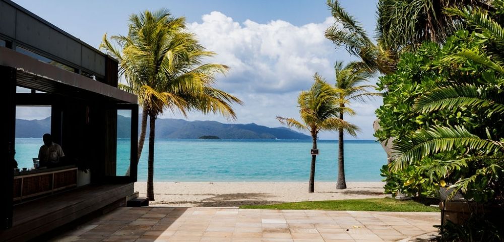 Tropical beach view with palm trees, turquoise waters, and distant mountains, framed by a modern building's patio area.