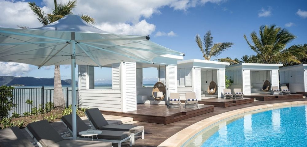 Relaxing poolside scene featuring lounge chairs, cabanas, and palm trees under a bright blue sky with fluffy clouds.
