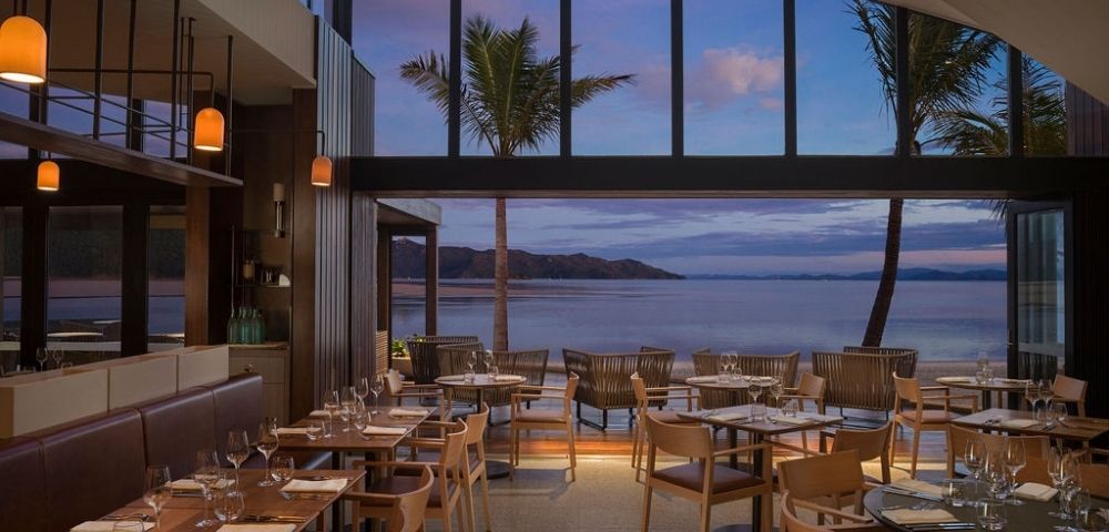 Chic restaurant interior with wooden tables and chairs, large windows framing a serene beach view at sunset, palm trees swaying outside.