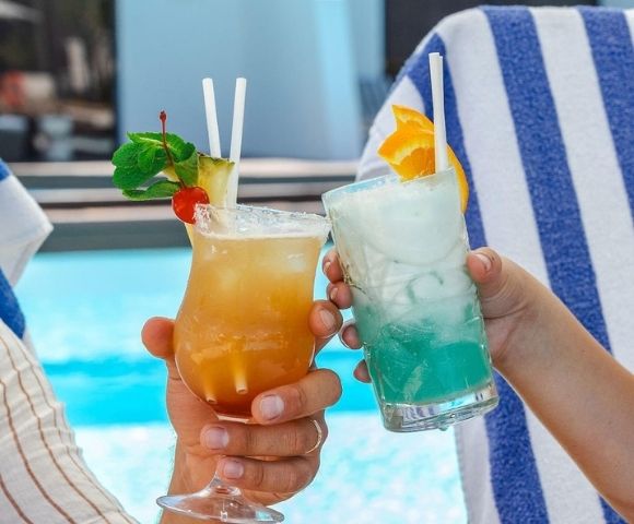 Two colorful cocktails are clinked together by hands, with a blue pool in the background, evoking a fun summer vibe.