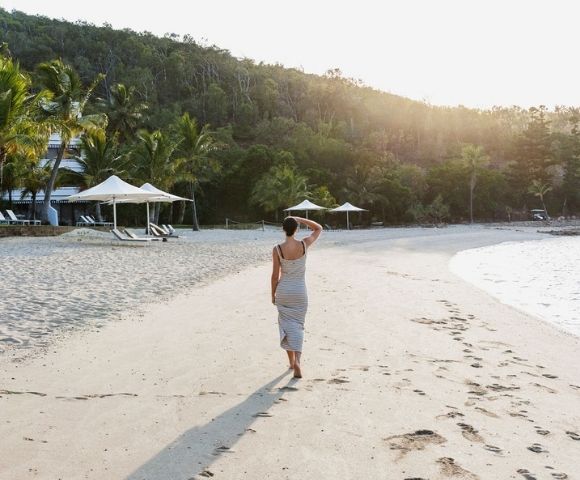 A woman in a flowing gray dress walks along a serene beach at sunset, palm trees and cabanas in the background.