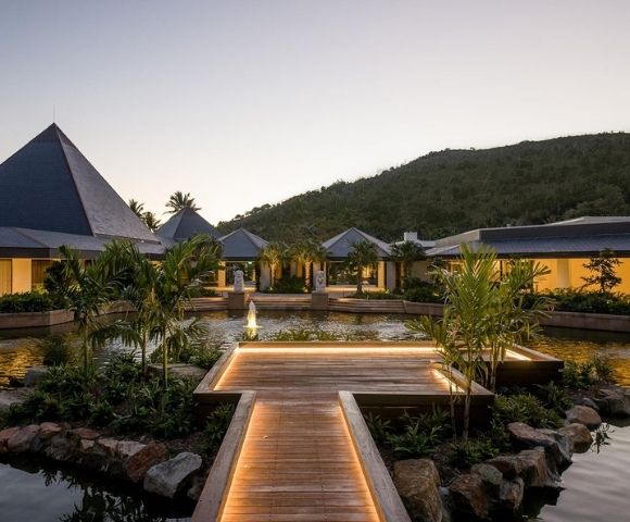 A serene resort at dusk, featuring a wooden walkway, lush greenery, and modern architecture set against a mountainous backdrop.