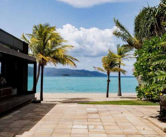 Tropical beach view with palm trees, turquoise waters, and distant mountains, framed by a modern building's patio area.