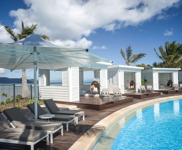 Relaxing poolside scene featuring lounge chairs, cabanas, and palm trees under a bright blue sky with fluffy clouds.