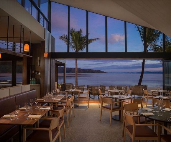 Chic restaurant interior with wooden tables and chairs, large windows framing a serene beach view at sunset, palm trees swaying outside.