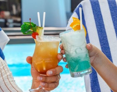 Two colorful cocktails are clinked together by hands, with a blue pool in the background, evoking a fun summer vibe.