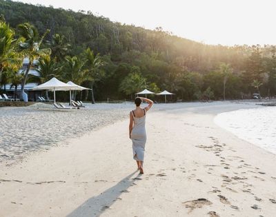 A woman in a flowing gray dress walks along a serene beach at sunset, palm trees and cabanas in the background.