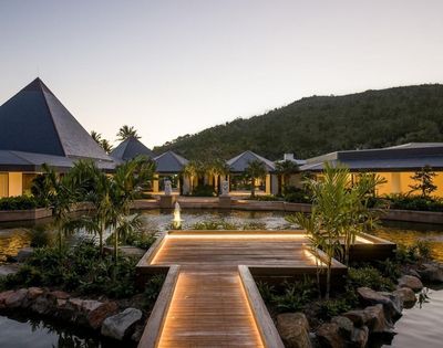 A serene resort at dusk, featuring a wooden walkway, lush greenery, and modern architecture set against a mountainous backdrop.