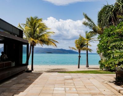 Tropical beach view with palm trees, turquoise waters, and distant mountains, framed by a modern building's patio area.