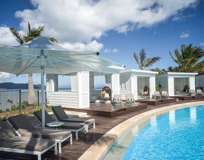 Relaxing poolside scene featuring lounge chairs, cabanas, and palm trees under a bright blue sky with fluffy clouds.