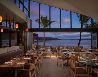 Chic restaurant interior with wooden tables and chairs, large windows framing a serene beach view at sunset, palm trees swaying outside.