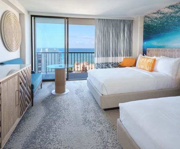 Bright hotel room with two beds, ocean view balcony, modern décor, and inviting seating area.