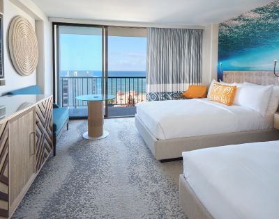 Bright hotel room with two beds, ocean view balcony, modern décor, and inviting seating area.
