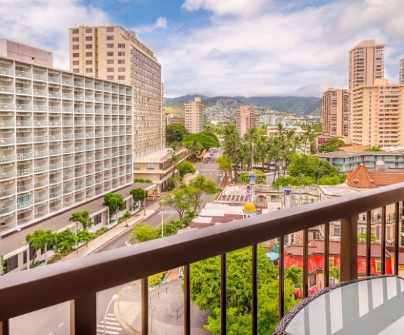 View from a balcony overlooking a vibrant city with palm trees, low-rise buildings, and mountains in the distance under a partly cloudy sky.