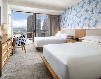 A cozy hotel room featuring two beds, a patterned chair, wooden furniture, and large windows with a city view.