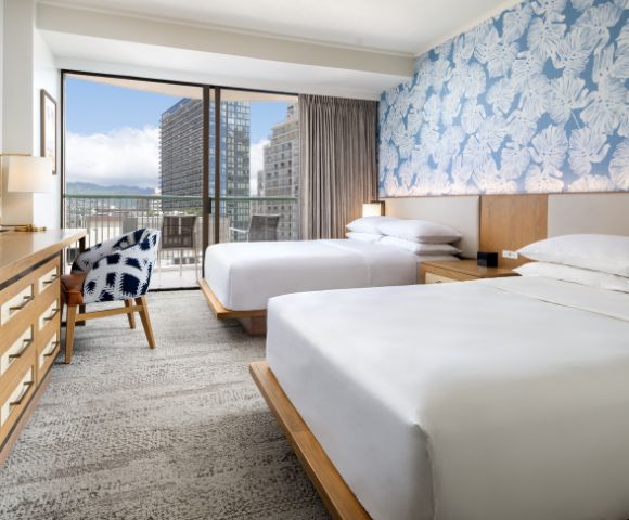 A cozy hotel room featuring two beds, a patterned chair, wooden furniture, and large windows with a city view.