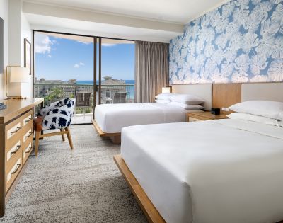Bright hotel room with two beds, coastal decor, a wooden dresser, and large window showcasing ocean views.