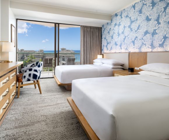 Bright hotel room with two beds, coastal decor, a wooden dresser, and large window showcasing ocean views.