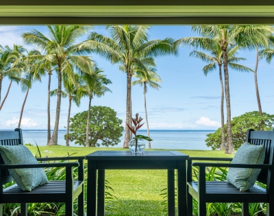 A serene view from a patio featuring two chairs and a table overlooking palm trees and a calm sea under a clear blue sky.