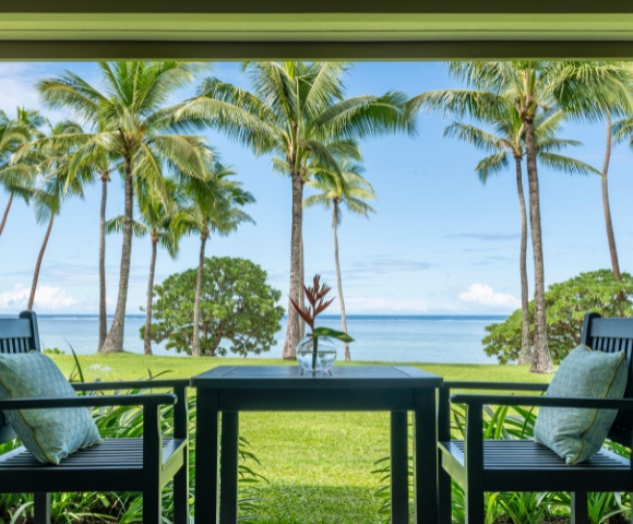 A serene view from a patio featuring two chairs and a table overlooking palm trees and a calm sea under a clear blue sky.